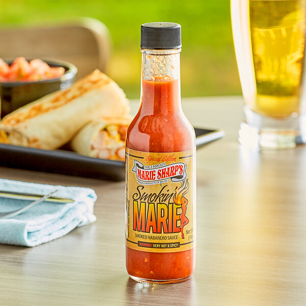 A bottle of Marie Sharp's Smokin' Marie Habanero hot sauce on a table with a bowl of food and a burrito.