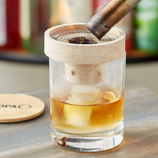A glass with a brown drink and ice being smoked with an Acopa Whiskey Smoker.