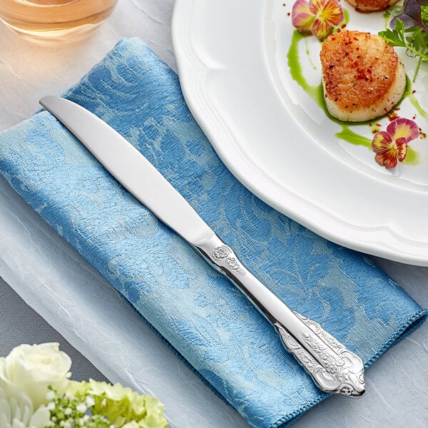 An Acopa Ophelia stainless steel dinner knife on a blue napkin next to a plate of scallops.
