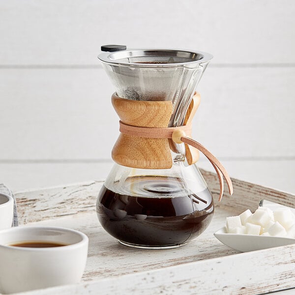 An Acopa glass coffee maker with a wood collar filled with brown liquid and a cup of coffee on a table.