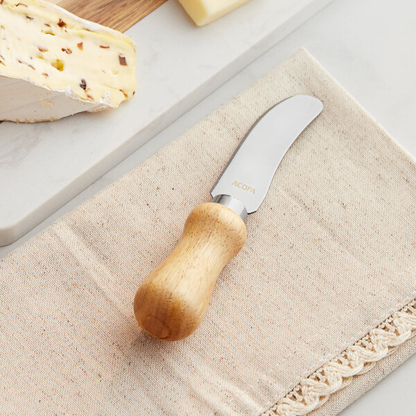 An Acopa stainless steel cheese spreader with a wood handle on a napkin next to cheese.