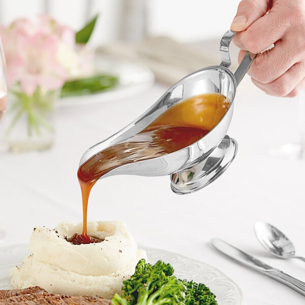 A person using a Choice stainless steel gravy boat to pour brown gravy over a plate of food.