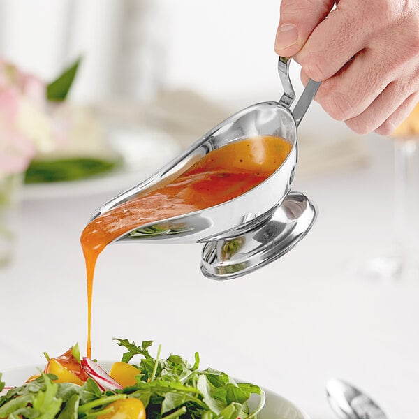 A hand pouring sauce from a stainless steel gravy boat into a bowl of salad.