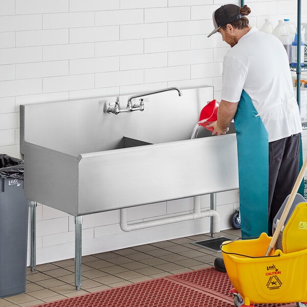 A man in a white shirt and blue apron cleaning a large stainless steel Regency utility sink.