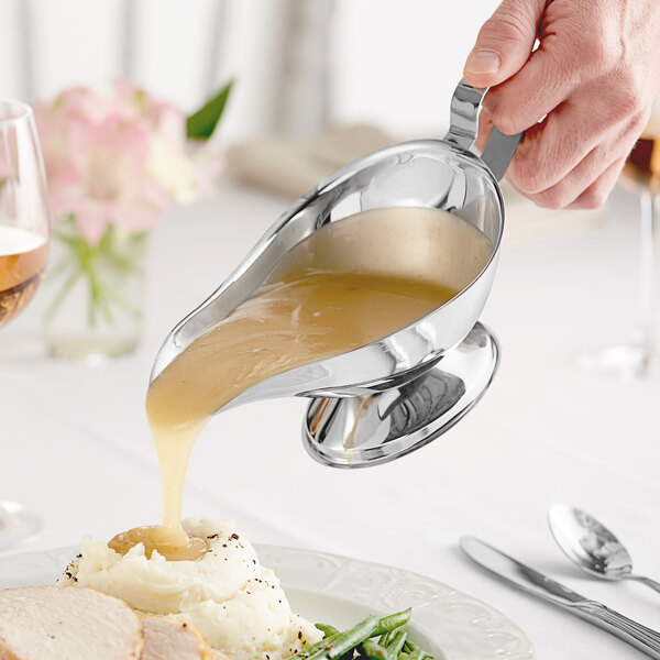 A person using a Choice stainless steel gravy boat to pour gravy over a plate of food.