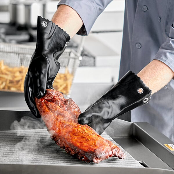A person holding a Mr. Bar-B-Q black oven glove over a piece of meat cooking on a grill.