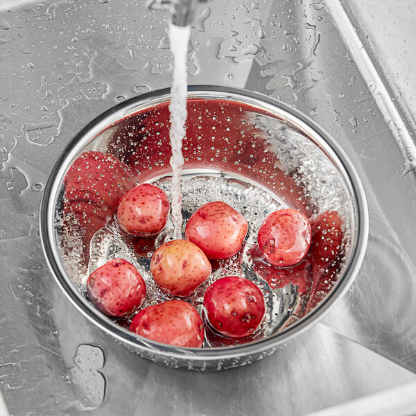 A stainless steel Chinese colander filled with red potatoes being washed under running water.