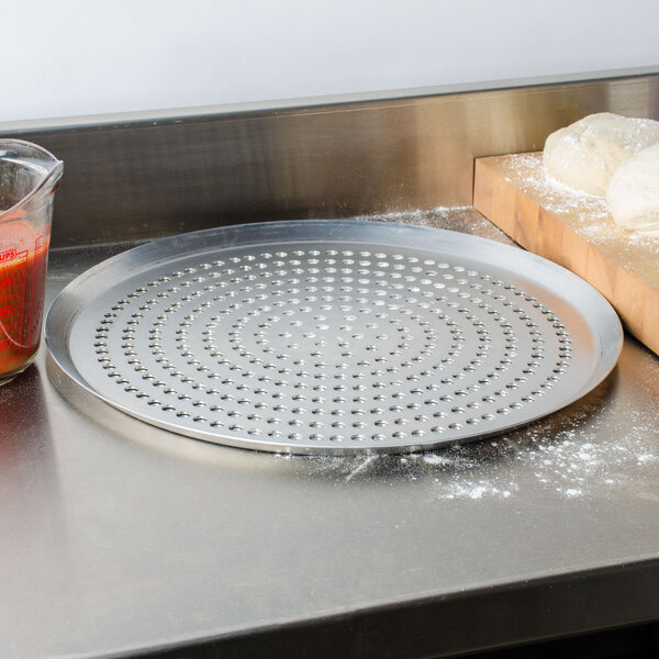 An American Metalcraft Super Perforated Heavy Weight Aluminum Pizza Pan with dough on it next to a cutting board and a container of liquid.