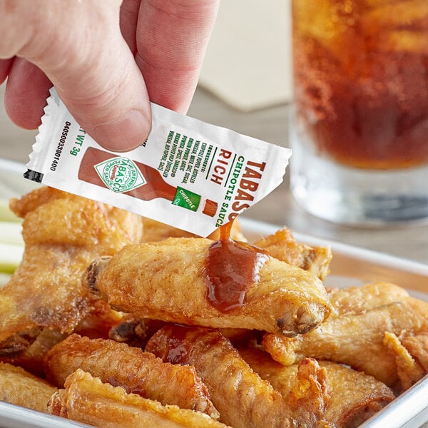 A hand holding a TABASCO Chipotle Hot Sauce packet over a tray of chicken wings.