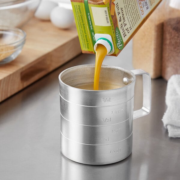 A person pouring orange juice from a carton into a Choice aluminum measuring cup.