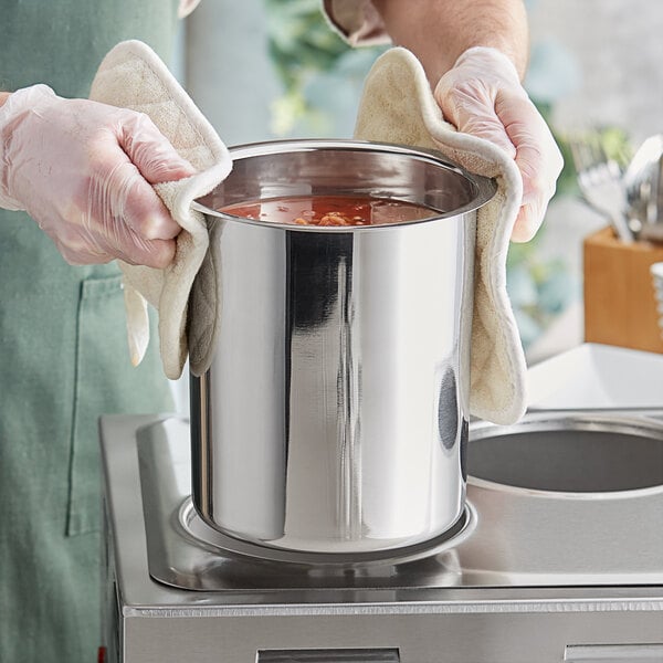 A person wearing gloves and an apron holding a Choice stainless steel bain marie pot filled with soup.