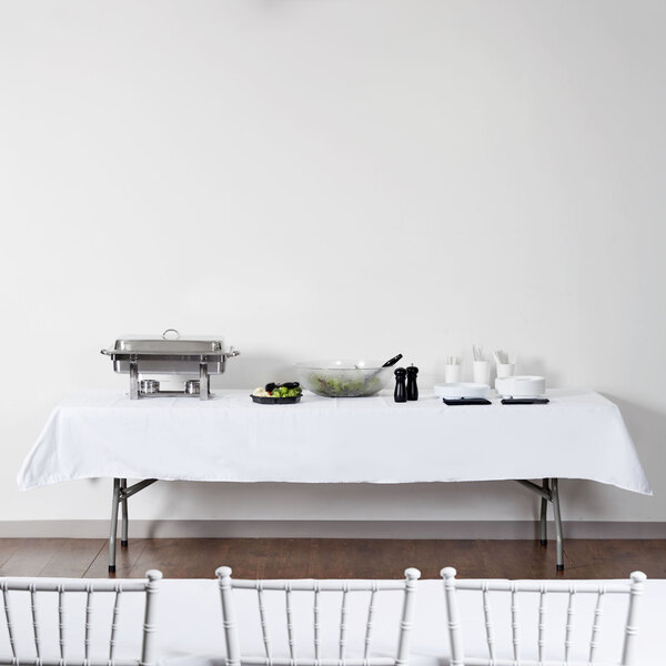 A rectangular white table with a white tablecloth and a bowl of vegetables on it.