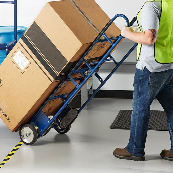 A man wearing a safety vest using a Lavex blue steel appliance hand truck to move a large box.