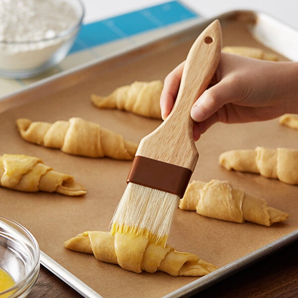 A person using a 2" wide boar bristle pastry brush with a wood handle to baste croissants on a tray.
