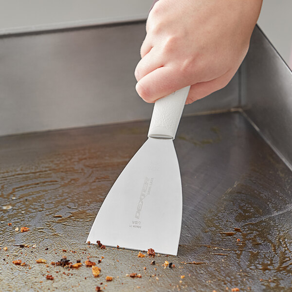 A hand using a Dexter-Russell grill scraper with a white plastic handle to clean a pan.