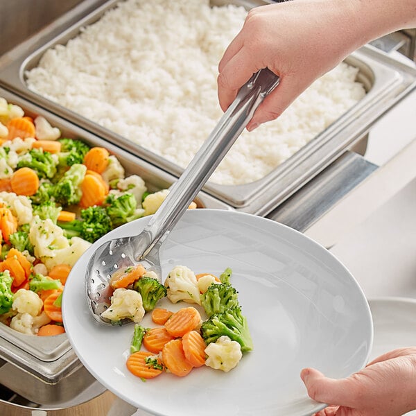 A hand holding a Choice stainless steel basting spoon over a plate of vegetables including broccoli and carrots.