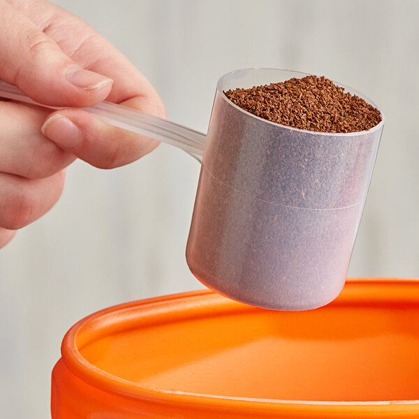 A hand using a long-handled polypropylene scoop to pour brown granules into a container.
