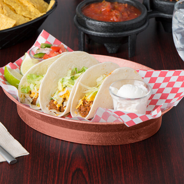 An oval deli server with a bowl of chips on a table with a plate of tacos and a red and white checkered napkin.
