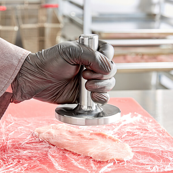 A person in black gloves using a round stainless steel meat tenderizer on a piece of meat.