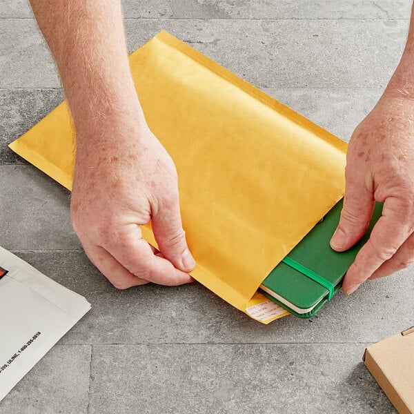 A hand opening a yellow Lavex Self-Sealing Kraft bubble mailer to reveal a green notebook.
