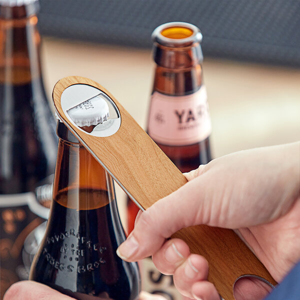 A person using a Franmara stainless steel bottle opener with wood overlay to open a bottle of beer.