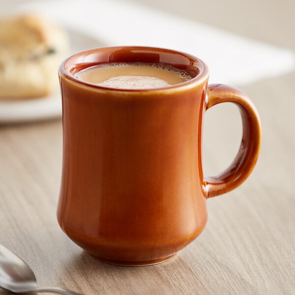 A brown Acopa stoneware coffee mug on a table with a spoon and a drink in it.