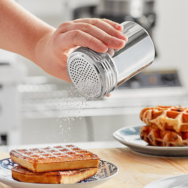 A hand holding a silver Choice stainless steel shaker sprinkling powder on a stack of waffles.