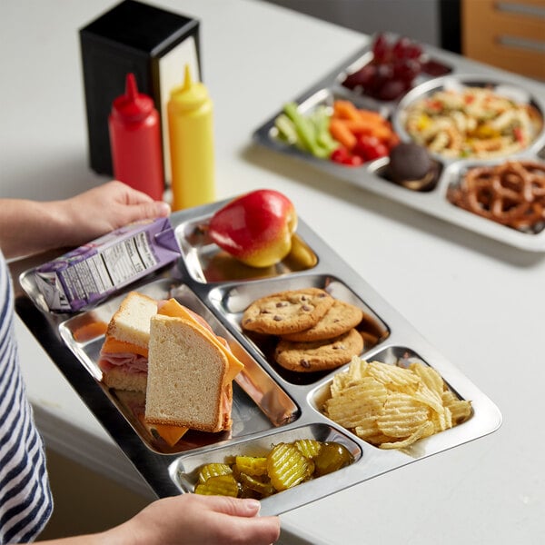 A person holding a Choice stainless steel rectangular tray with 6 compartments filled with food.