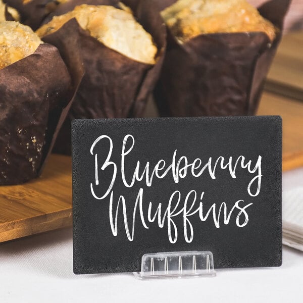 A brown table with blueberry muffins and a black sign with white text that reads "Blueberry Muffins" next to a brown paper sign holder.