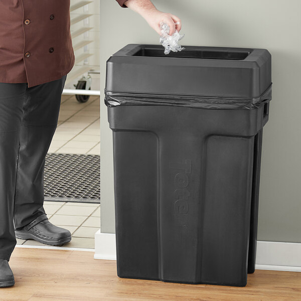 A man in a chef's outfit standing next to a Toter black trash can with a black lid.