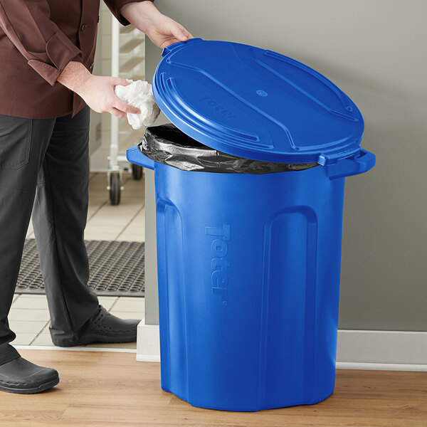 A person putting a plastic bag in a blue Toter trash can.