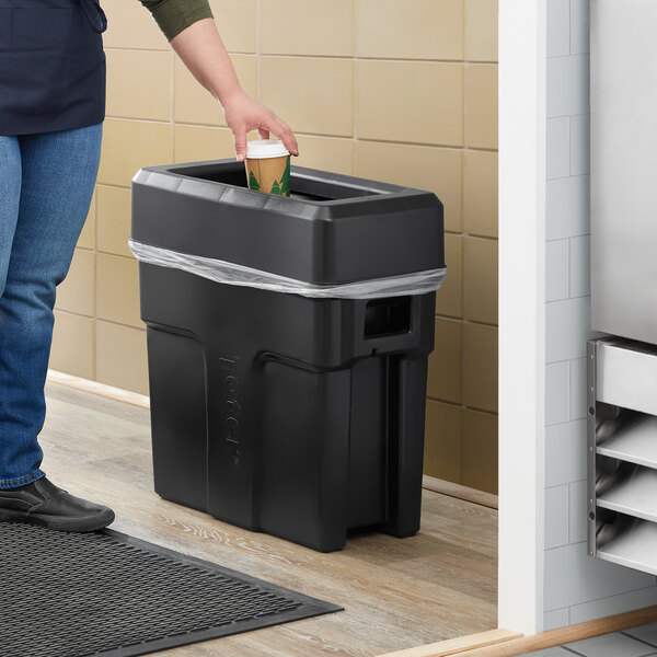 A woman pouring a drink into a Toter Slimline black trash can in a school kitchen.