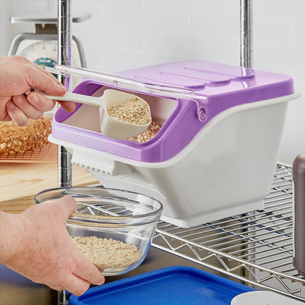 A person using a scoop to pour oats into a Baker's Mark purple ingredient bin.