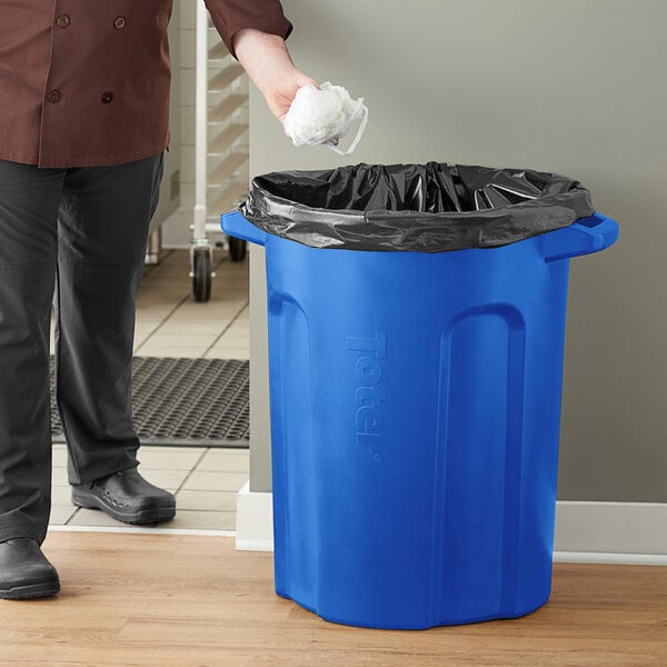 A man in a chef's uniform standing next to a Toter blue trash can with a black bag.