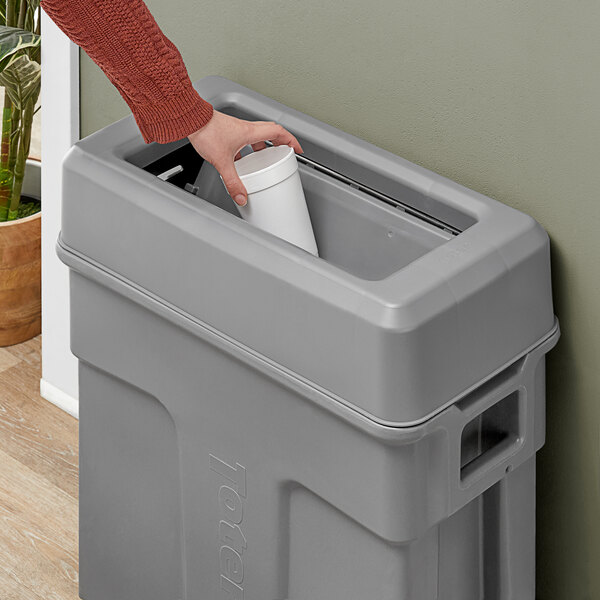 A hand using a Toter dark cool gray swing lid to put a white container in a trash can.