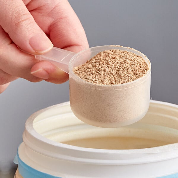 A hand using a 43 cc polypropylene scoop to pour protein powder into a plastic container.