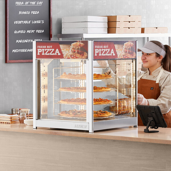 A woman standing behind a ServIt countertop pizza warmer with pizzas in a glass case.