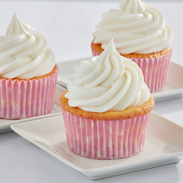 Three Enjay pink fluted baking cups with white polka dots holding cupcakes with white frosting.