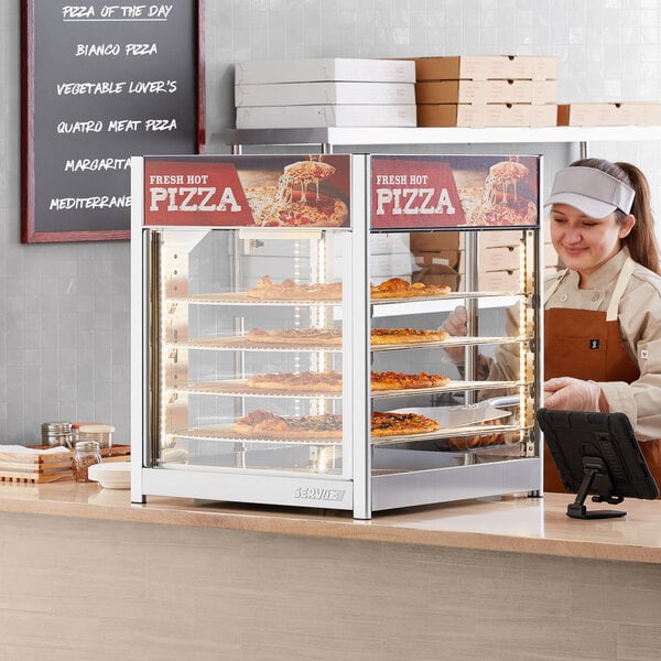 A woman wearing a white cap and apron standing behind a ServIt countertop display warmer filled with pizzas.