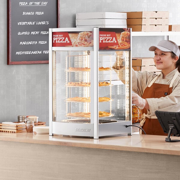 A woman in a white apron standing behind a ServIt pizza warmer display case with a pizza.