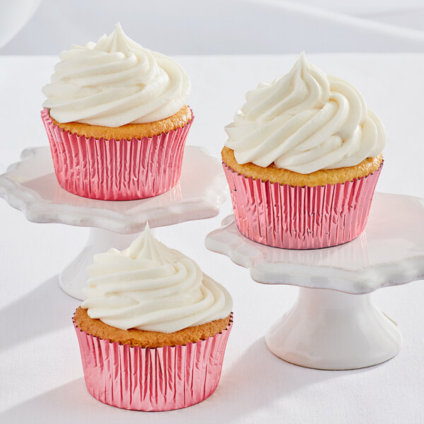 Three Enjay pink foil baking cups with white frosted cupcakes on white pedestals.