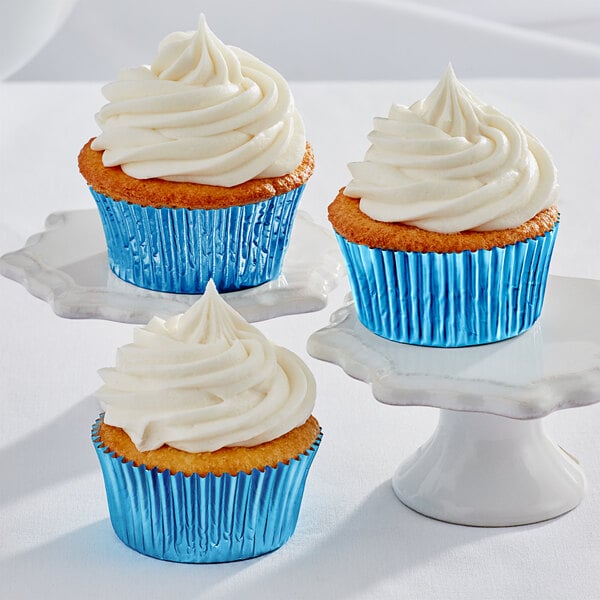 Three Enjay blue foil baking cups with cupcakes with white frosting on a white pedestal.