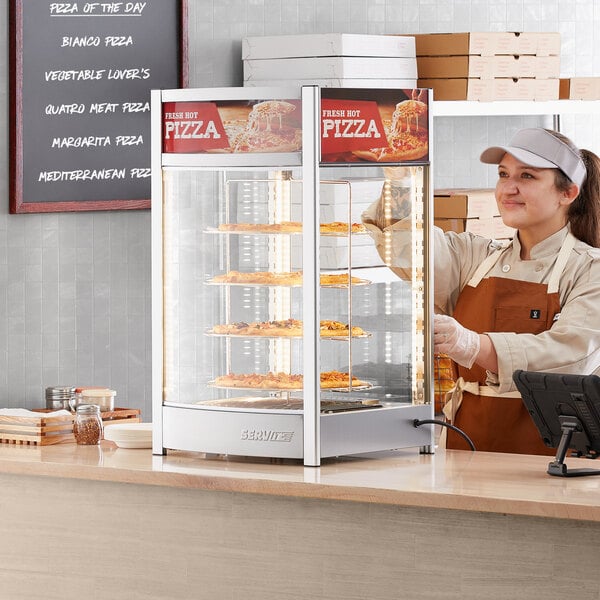 A woman wearing an apron standing behind a ServIt pizza warmer display case with a pizza inside.