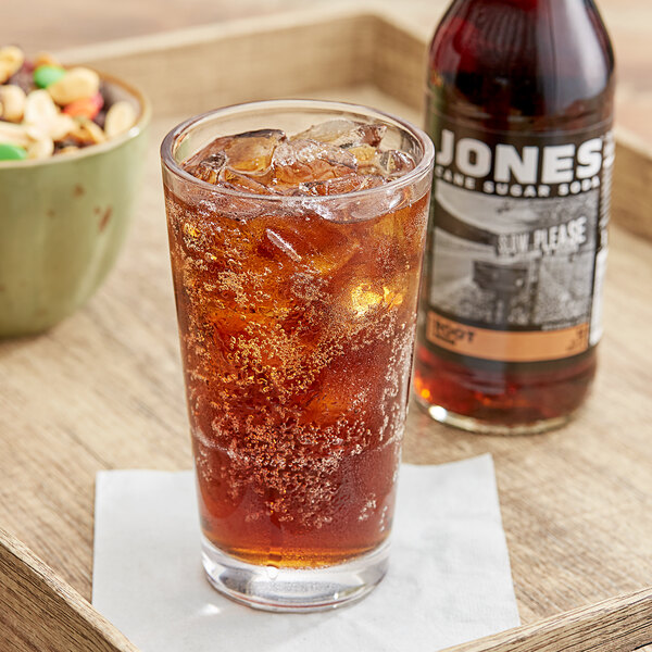 A glass of Jones Root Beer soda with ice on a table next to a bottle of nuts.