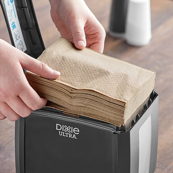 A person putting a Dixie Ultra Brown interfold paper napkin into a dispenser on a counter in a professional kitchen.