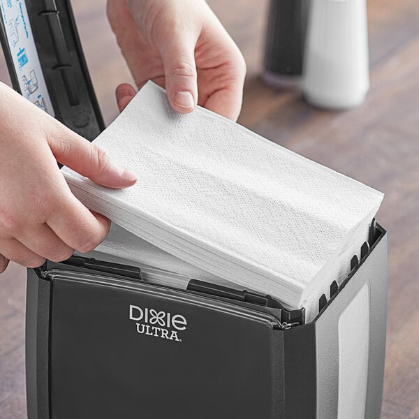 A person putting a Dixie Ultra white interfold paper napkin into a dispenser on a counter in a professional kitchen.