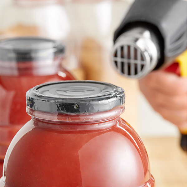A hand using a yellow and red sprayer to apply a clear shrink band to a jar of red sauce.