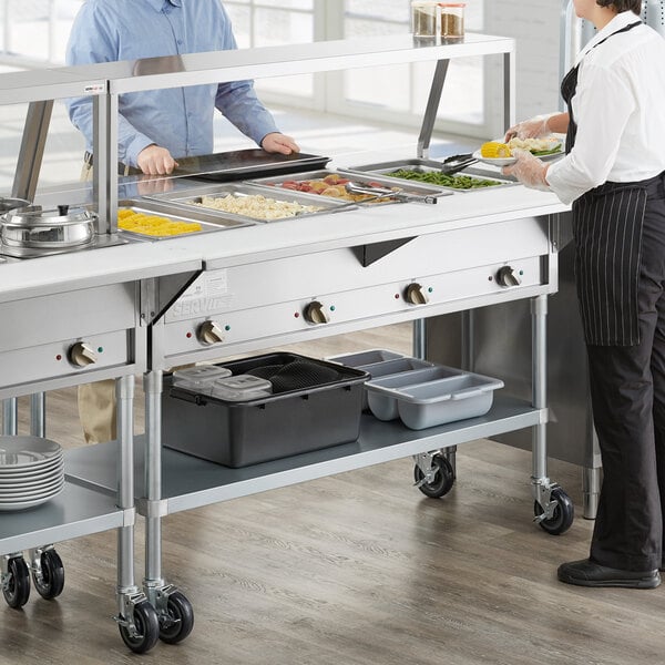A man and woman using a ServIt electric steam table with sneeze guard in a school kitchen.
