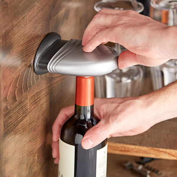 A person using a Franmara silver wall-mount foil cutter to open a bottle of wine.