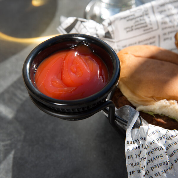 A close up of a Thunder Group black fluted melamine ramekin filled with ketchup on a hamburger.